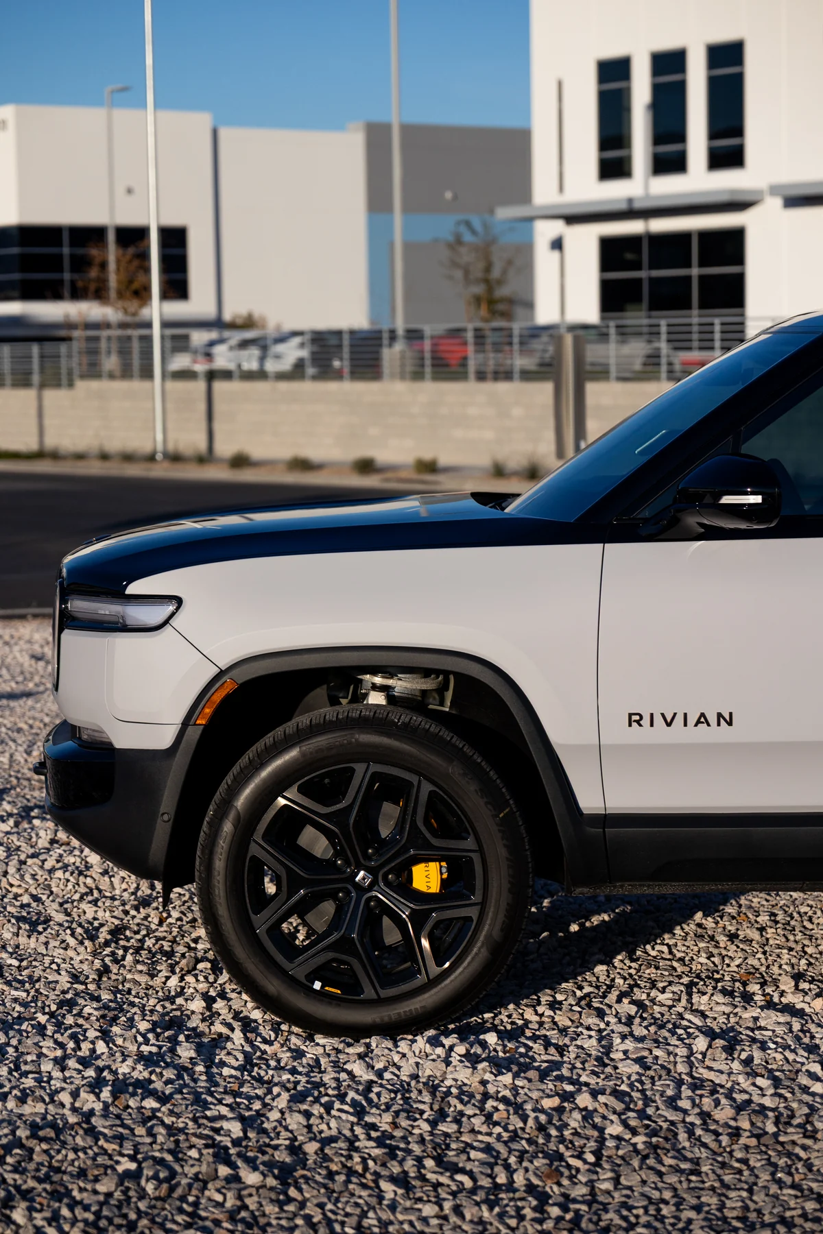 Edited photograph of a two-tone white and dark blue Rivian R1S, front quarter panel and wheel detail, shot on gravel outside a commercial building