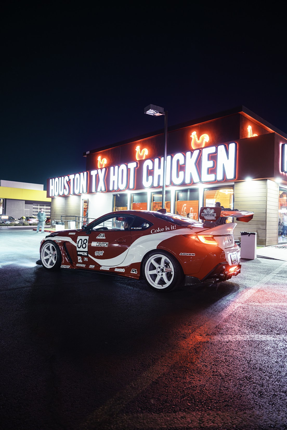 Orange and white liveried Toyota GR86 with a big rear wing parked outside a neon lit Las Vegas storefront at night during SEMA week