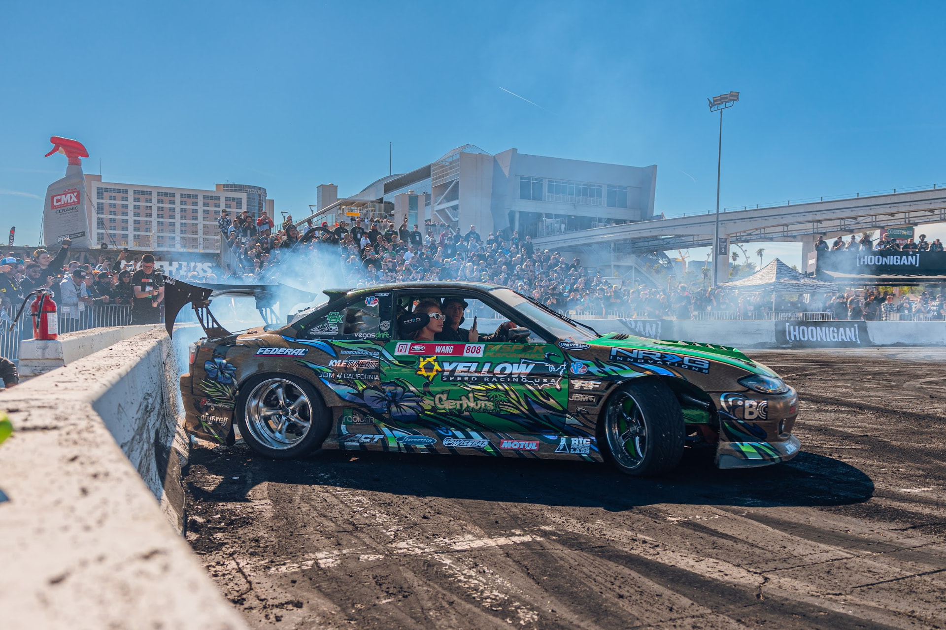 Green and gold Nissan S15 drift car sliding with a ride along passenger in front of the SEMA Ignited grandstand crowd