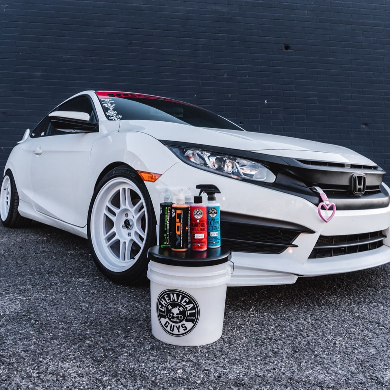 White Honda Civic coupe on white wheels staged with detailing products on a bucket against a dark brick wall